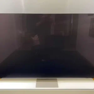 Der PHOTO-2024-06-21-12-10-062-1, ein 85 Zoll großer Samsung 8K-Fernseher, ist ausgeschaltet und steht auf einem Ständer in einem hell erleuchteten Raum mit gelb-weißen Wänden. Auf dem Bildschirm sind Reflexionen des gut beleuchteten Raumes sichtbar.